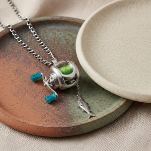 Pode incluir: Um colar com pingente de carretel de pesca prateado com linha de pesca verde e detalhes turquesa. Um pequeno pingente de peixe pende abaixo do carretel. O colar está em uma corrente.