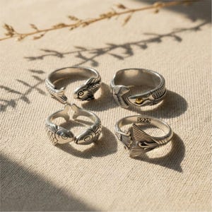 Puede incluir: Cuatro anillos de tono plateado con motivos de peces. Dos anillos muestran cabezas de peces que se encuentran, un anillo muestra un pez con un pequeño acento dorado y un anillo presenta un diseño detallado de cola de pez con un sello S925.