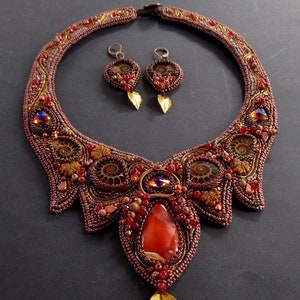 May include: A beaded necklace and earrings set with a large carnelian stone pendant. The necklace is made with brown, red, and gold beads and features a swirling pattern. The earrings are small and feature a similar design.
