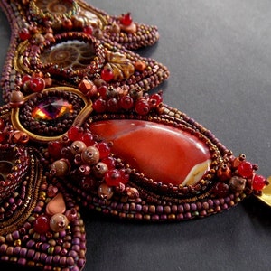May include: A close-up of a beaded necklace with a large red gemstone pendant. The necklace is made with a variety of beads, including brown, gold, and red. The pendant is surrounded by smaller red beads and a gold leaf.