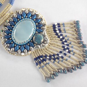 Puede incluir: Un collar de cuentas con un colgante azul y plateado con una piedra ovalada azul claro. El colgante tiene una franja de cuentas azules y doradas.
