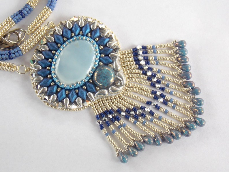 Puede incluir: Un collar de cuentas con un colgante azul y plateado con una piedra ovalada azul claro. El colgante tiene una franja de cuentas azules y doradas.
