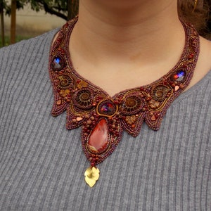 May include: A statement necklace with a copper and gold color scheme. The necklace features a large, red gemstone pendant and intricate beadwork.