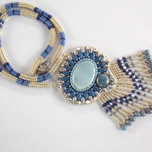 Puede incluir: Un collar de cuentas con una cadena azul y dorada y un gran colgante con un dise&ntilde;o de cuentas azules y plateadas con una piedra ovalada azul claro en el centro.