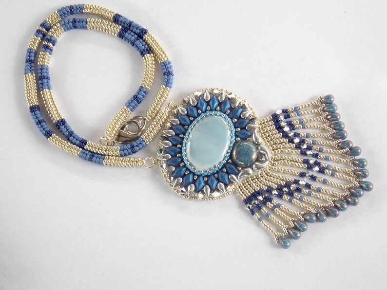 Puede incluir: Un collar de cuentas con una cadena azul y dorada y un gran colgante con un dise&ntilde;o de cuentas azules y plateadas con una piedra ovalada azul claro en el centro.