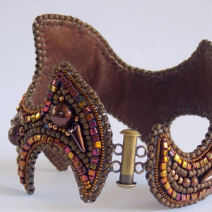May include: A handcrafted brown beaded cuff bracelet with a leather backing. The bracelet features a complex design with various sizes and colours of beads, including bronze, gold, and iridescent accents. It has a metal clasp closure.