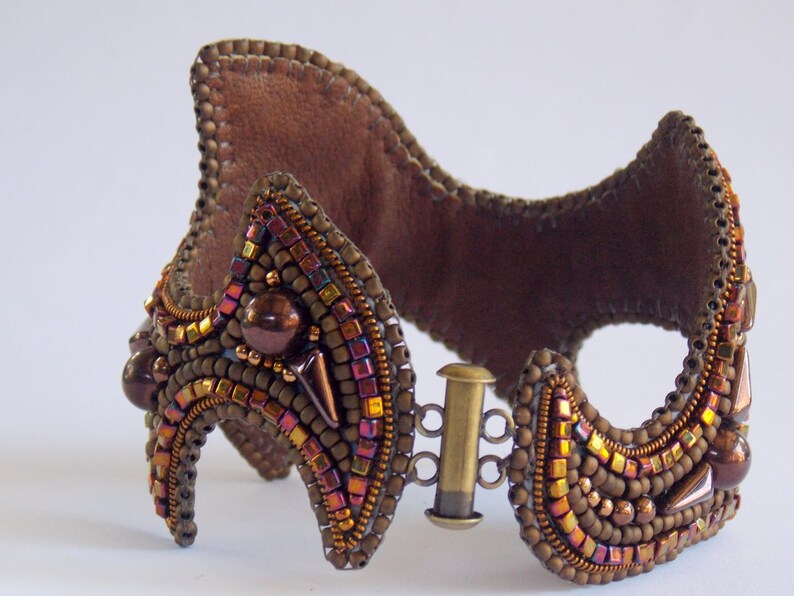 May include: A handcrafted brown beaded cuff bracelet with a leather backing. The bracelet features a complex design with various sizes and colours of beads, including bronze, gold, and iridescent accents. It has a metal clasp closure.
