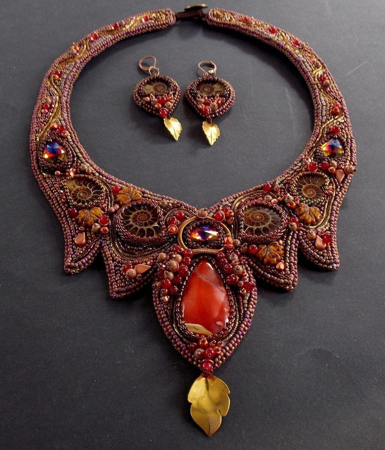 May include: A beaded necklace and earrings set with a large carnelian stone pendant. The necklace is made with brown, red, and gold beads and features a swirling pattern. The earrings are small and feature a similar design.