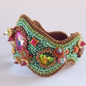 May include: A beaded bracelet featuring a green, brown and gold colour scheme. The bracelet is adorned with a variety of colourful crystals and beads.