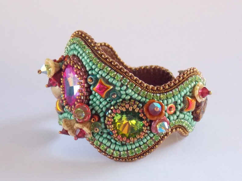 May include: A beaded bracelet featuring a green, brown and gold colour scheme. The bracelet is adorned with a variety of colourful crystals and beads.