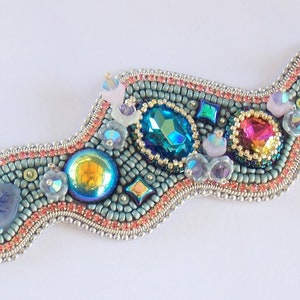 May include: A beaded bracelet with a wavy design features a variety of colourful gemstones, including turquoise, pink, and iridescent blue. The bracelet is made with silver-toned metal clasps.