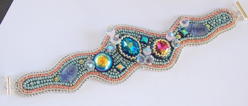 May include: A beaded bracelet with a wavy design features a variety of colourful gemstones, including turquoise, pink, and iridescent blue. The bracelet is made with silver-toned metal clasps.