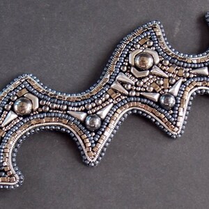 May include: A beaded bracelet with a distinctive, undulating shape. The bracelet combines silver, grey, and blue beads, with metallic highlights and a secure clasp. The detailed beadwork creates a textured, striking accessory, suitable for any occasion.