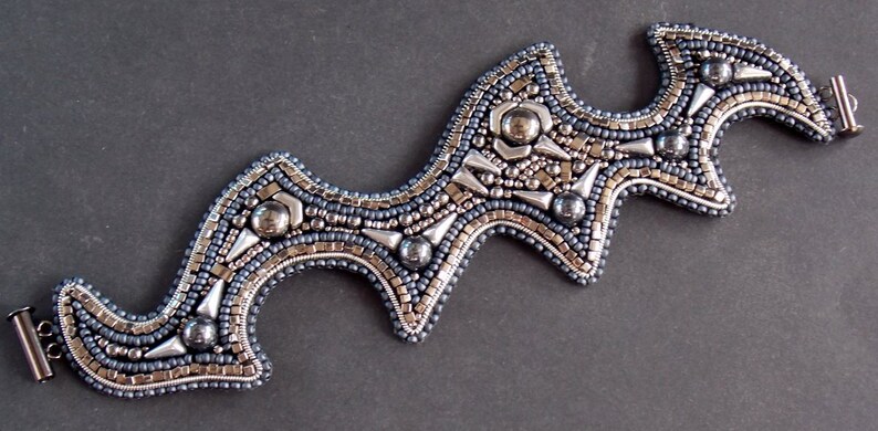 May include: A beaded bracelet with a distinctive, undulating shape. The bracelet combines silver, grey, and blue beads, with metallic highlights and a secure clasp. The detailed beadwork creates a textured, striking accessory, suitable for any occasion.