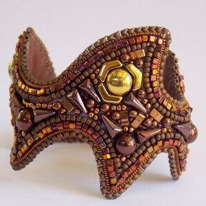 May include: A handcrafted cuff bracelet with a mosaic of small, iridescent beads in shades of brown, gold, and copper. The central design includes a gold-coloured metal accent. The bracelet has a unique, angular shape.