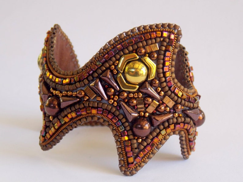 May include: A handcrafted cuff bracelet with a mosaic of small, iridescent beads in shades of brown, gold, and copper. The central design includes a gold-coloured metal accent. The bracelet has a unique, angular shape.