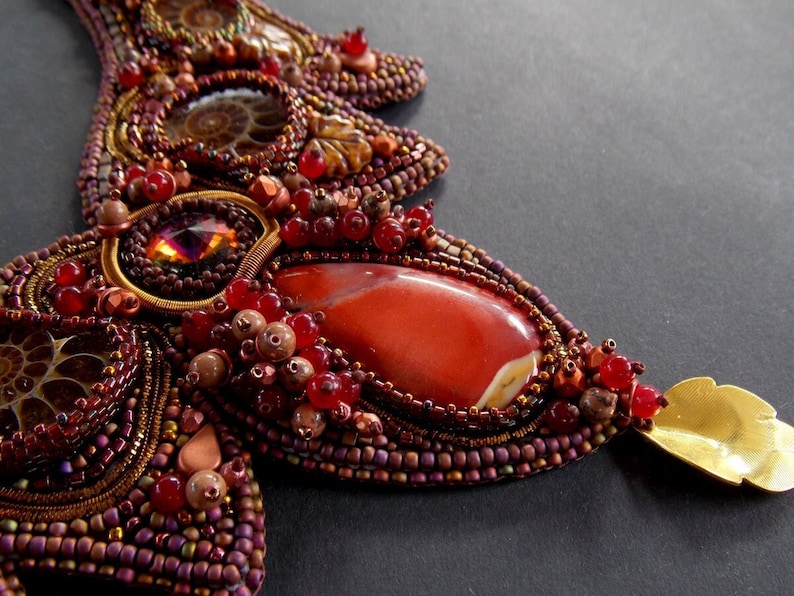 May include: A close-up of a beaded necklace with a large red gemstone pendant. The necklace is made with a variety of beads, including brown, gold, and red. The pendant is surrounded by smaller red beads and a gold leaf.