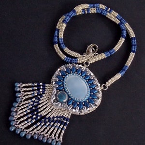 Puede incluir: Un collar de cuentas plateadas y azules con un gran colgante de forma ovalada. El colgante est&aacute; hecho de cuentas plateadas y azules y presenta una piedra azul claro en el centro. El collar mide aproximadamente 50 cm de largo.