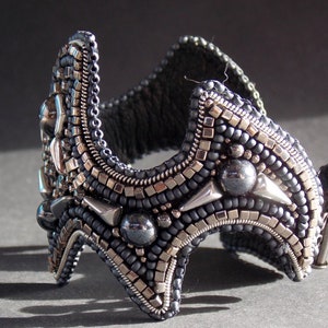 May include: A detailed cuff bracelet, crafted with dark grey and silver beads. The bracelet has a curved form with geometric details and a clasp. The intricate beadwork creates a textured, striking accessory, suitable for a special occasion.