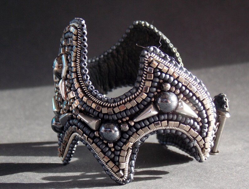 May include: A detailed cuff bracelet, crafted with dark grey and silver beads. The bracelet has a curved form with geometric details and a clasp. The intricate beadwork creates a textured, striking accessory, suitable for a special occasion.