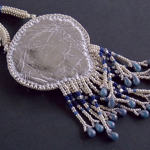 Puede incluir: Un collar con un colgante de plata, de forma ovalada y texturado. El colgante est&aacute; rodeado de una franja de cuentas plateadas y azules.