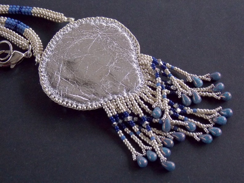 Puede incluir: Un collar con un colgante de plata, de forma ovalada y texturado. El colgante est&aacute; rodeado de una franja de cuentas plateadas y azules.