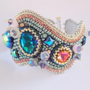 May include: A beaded bracelet with a wavy design, featuring a large teal blue gemstone, iridescent stones, and pink and purple accents.