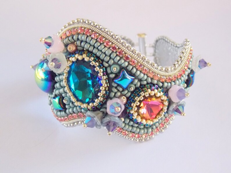 May include: A beaded bracelet with a wavy design, featuring a large teal blue gemstone, iridescent stones, and pink and purple accents.