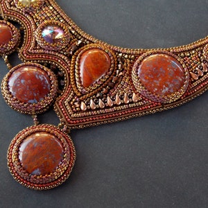 May include: A beaded necklace with a copper and brown color scheme. The necklace features several large, round, red and brown stones with a mottled pattern. The stones are surrounded by smaller beads and copper accents.
