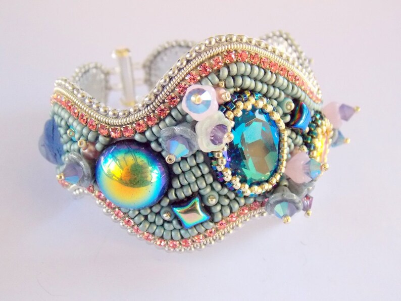 May include: A beaded bracelet with a wavy design, featuring a variety of colourful beads, including iridescent, blue, pink, and gold. The bracelet is adorned with a large, faceted, teal gemstone.