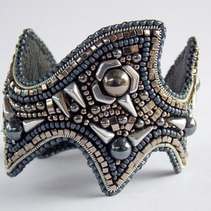 May include: A handcrafted, grey and silver beaded cuff bracelet. The bracelet has a crown-like design with metallic accents, including silver spikes and a central flower-shaped embellishment. The intricate beadwork creates a textured accessory.