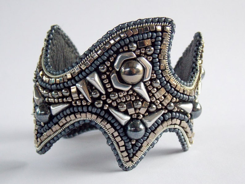 May include: A handcrafted, grey and silver beaded cuff bracelet. The bracelet has a crown-like design with metallic accents, including silver spikes and a central flower-shaped embellishment. The intricate beadwork creates a textured accessory.
