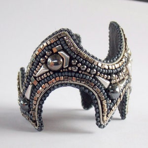 May include: A handcrafted cuff bracelet with a dark grey base, adorned with silver and metallic beads. The bracelet features geometric shapes and a curved design. The cuff is approximately 5 cm wide.