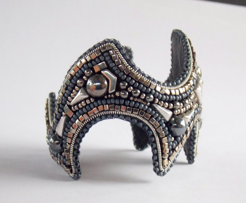 May include: A handcrafted cuff bracelet with a dark grey base, adorned with silver and metallic beads. The bracelet features geometric shapes and a curved design. The cuff is approximately 5 cm wide.