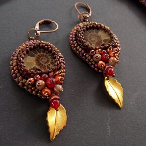May include: A pair of earrings with a brown and gold leaf design. The earrings feature a round, brown bead with a fossilized ammonite in the center. The beads are surrounded by smaller, brown, gold, and red beads.