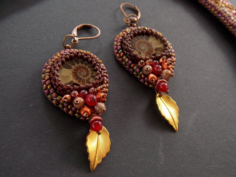 May include: A pair of earrings with a brown and gold leaf design. The earrings feature a round, brown bead with a fossilized ammonite in the center. The beads are surrounded by smaller, brown, gold, and red beads.