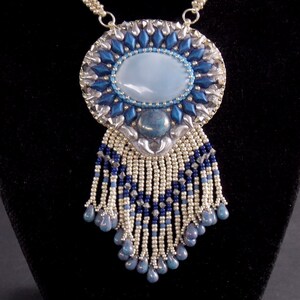 Puede incluir: Un collar de cuentas plateadas y azules con un gran colgante ovalado que presenta una piedra azul claro y una franja de cuentas.