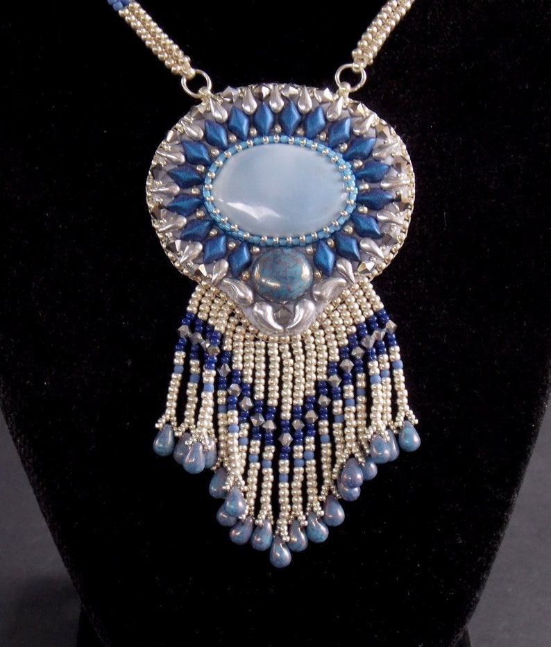Puede incluir: Un collar de cuentas plateadas y azules con un gran colgante ovalado que presenta una piedra azul claro y una franja de cuentas.