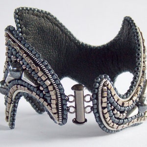 May include: A handcrafted cuff bracelet with a dark grey leather backing and detailed beadwork. The bracelet features silver and grey beads, metallic accents, and a silver clasp. The design has a unique, architectural shape.