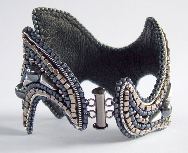 May include: A handcrafted cuff bracelet with a dark grey leather backing and detailed beadwork. The bracelet features silver and grey beads, metallic accents, and a silver clasp. The design has a unique, architectural shape.