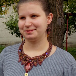 May include: A woman wearing a gray sweater with a large, intricate necklace made of brown, red, and gold beads. The necklace features a large, red gemstone pendant.