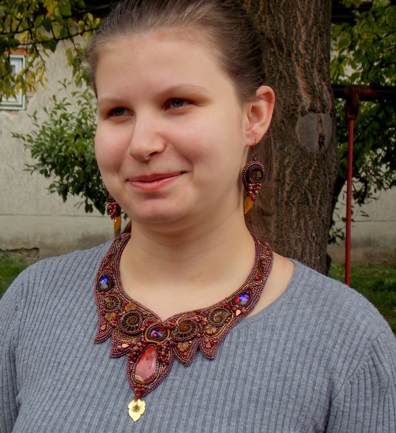 May include: A woman wearing a gray sweater with a large, intricate necklace made of brown, red, and gold beads. The necklace features a large, red gemstone pendant.
