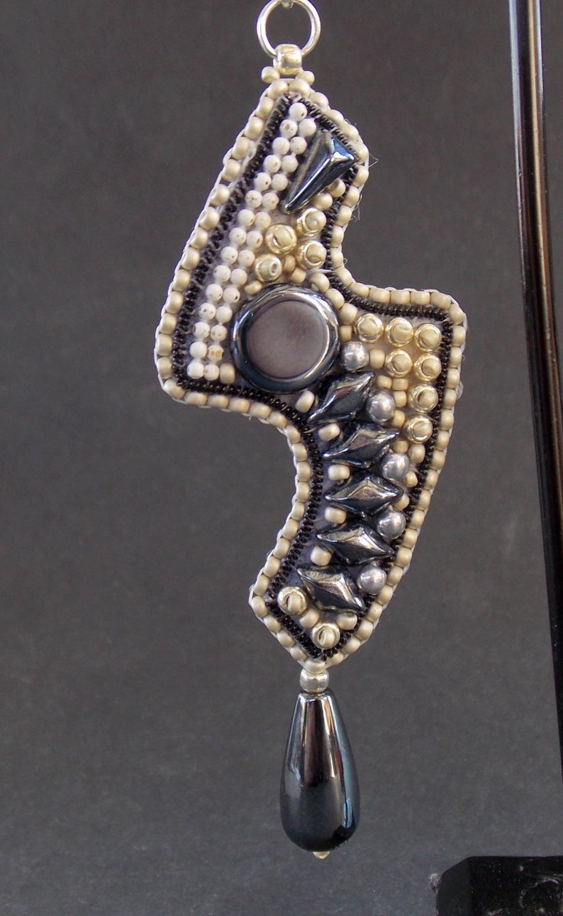Pode incluir: Um pingente de contas prateadas e pretas com uma pedra preta em forma de l&aacute;grima pendurada nele. O pingente tem a forma de um raio e tem uma pedra redonda cinza no centro.
