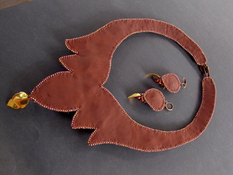 May include: A brown leather necklace with a gold pendant and matching earrings. The necklace is decorated with small beads and the earrings have a teardrop shape.