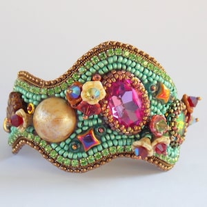 May include: A green, pink and gold beaded bracelet with a large pink gemstone and a pearl-like bead. The bracelet is made of small beads and has a wavy shape.