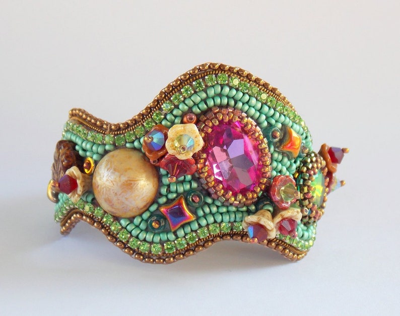 May include: A green, pink and gold beaded bracelet with a large pink gemstone and a pearl-like bead. The bracelet is made of small beads and has a wavy shape.