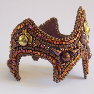 May include: A handcrafted beaded cuff bracelet in shades of brown, copper, and gold. The intricate design features various bead sizes and shapes, with metallic accents and a unique, architectural silhouette. The bracelet is approximately 15 cm in length.
