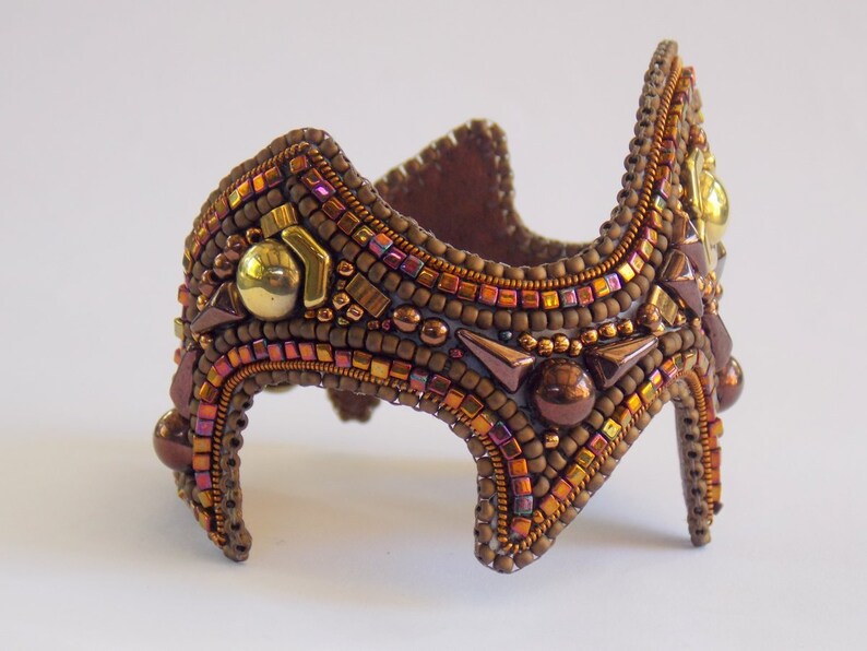 May include: A handcrafted beaded cuff bracelet in shades of brown, copper, and gold. The intricate design features various bead sizes and shapes, with metallic accents and a unique, architectural silhouette. The bracelet is approximately 15 cm in length.