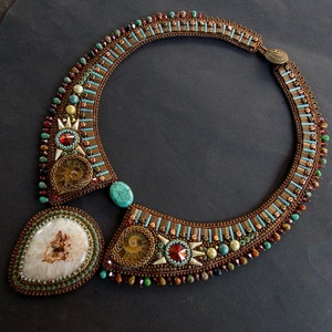 May include: A beaded collar necklace featuring a large white geode pendant, two spiral ammonite fossils, turquoise cabochons, and red faceted crystals. The piece is intricately embroidered with brown, teal, and metallic beads.