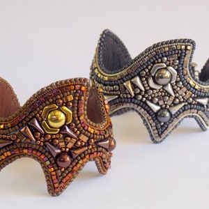 May include: Two beaded cuff bracelets. One is bronze and gold, with a central gold accent. The other is silver and grey, with silver spikes and a central silver accent. Both have a curved shape and a leather backing.